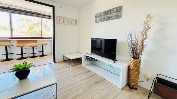 Een moderne woonkamer met een televisie, een glazen salontafel, een plant en een zitgedeelte bij het raam.