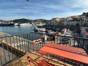 Een balkon met een tafel en stoelen, met uitzicht op een haven vol boten en gebouwen.