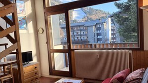 Ein Zimmer mit großem Fenster, einem Holztisch, einem Bett mit Kissen, einem Heizkörper und einem Fernseher auf einem Holzständer.