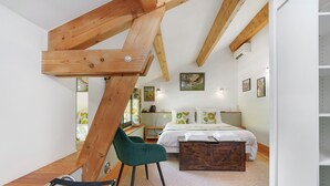 Ein Schlafzimmer mit schrägen Wänden, Holzbalken, einem Bett, einem Schreibtisch, einem Stuhl und Kunstwerken an den Wänden.