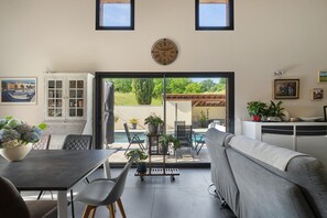 A modern living room with a dining area, a sofa, a coffee table, and a view of a pool and greenery.