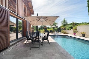 Un espace aménagé au bord de la piscine, avec une table et des chaises, un grand parasol et une piscine.