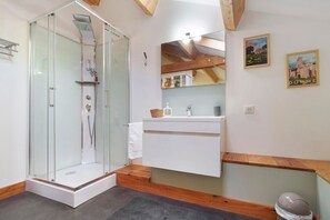 Une salle de bain moderne avec une douche à l’italienne en verre, un meuble-lavabo blanc avec un miroir et des poutres en bois au plafond.