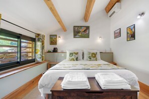 Une chambre à coucher avec un lit, des oreillers, une table de chevet, une fenêtre avec des rideaux et des œuvres d’art accrochées aux murs.