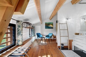 Quarto com vigas de madeira, janela com cortinas, um quadro, duas cadeiras, uma escada e um aparador branco.