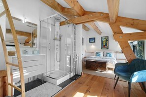 A modern bedroom with a wooden ceiling, a glass shower, a bed with blue bedding, and a blue chair.