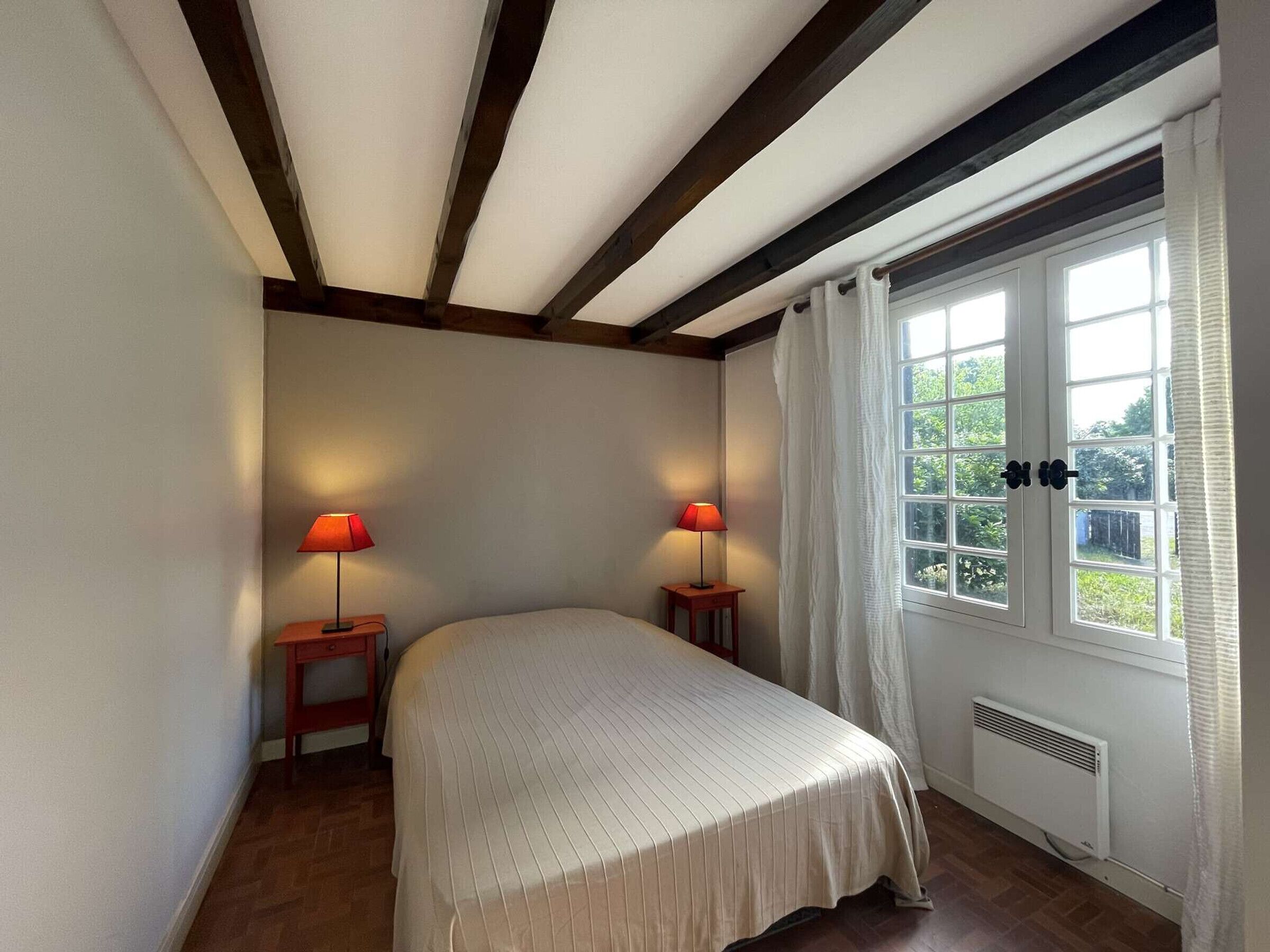 A bedroom with a bed, two bedside lamps, a window with curtains, and a radiator.
