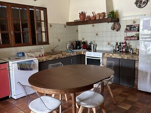 Eine kleine Küche mit rundem Tisch, Stühlen, einem Kühlschrank, einem Herd und einem Fenster mit Holzläden.