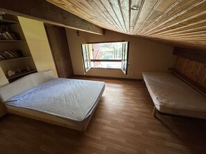 Ein kleines, einzelnes Zimmer mit Holzboden, einem Bett, einer Bank und einem Fenster mit Blick ins Freie.