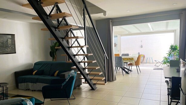 A modern living room with a staircase, a sofa, a dining area, and a view through a glass door. - Modern villa with pool and parking - La Grande Motte (La Grande-Motte)