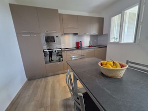 A modern kitchen with built-in appliances, a dining table, and a window with blinds. - Apartment with air conditioning, terrace - 3 guests (Lumio)
