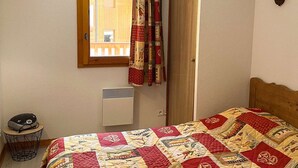 A bedroom with a wooden headboard, a quilted bedspread, a window with curtains, a wall-mounted lamp, and a radiator. - Comfortable and bright apartment - 4 guests (Valloire)