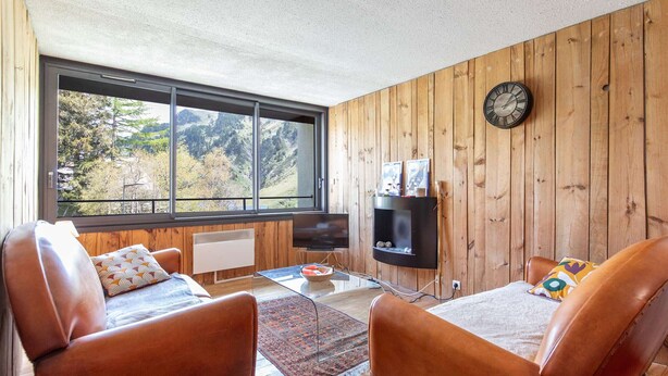Une pièce comprenant deux canapés en cuir, une table basse en verre, un téléviseur posé sur un meuble, un mur en bois orné d’une horloge, et une grande fenêtre donnant sur la verdure.