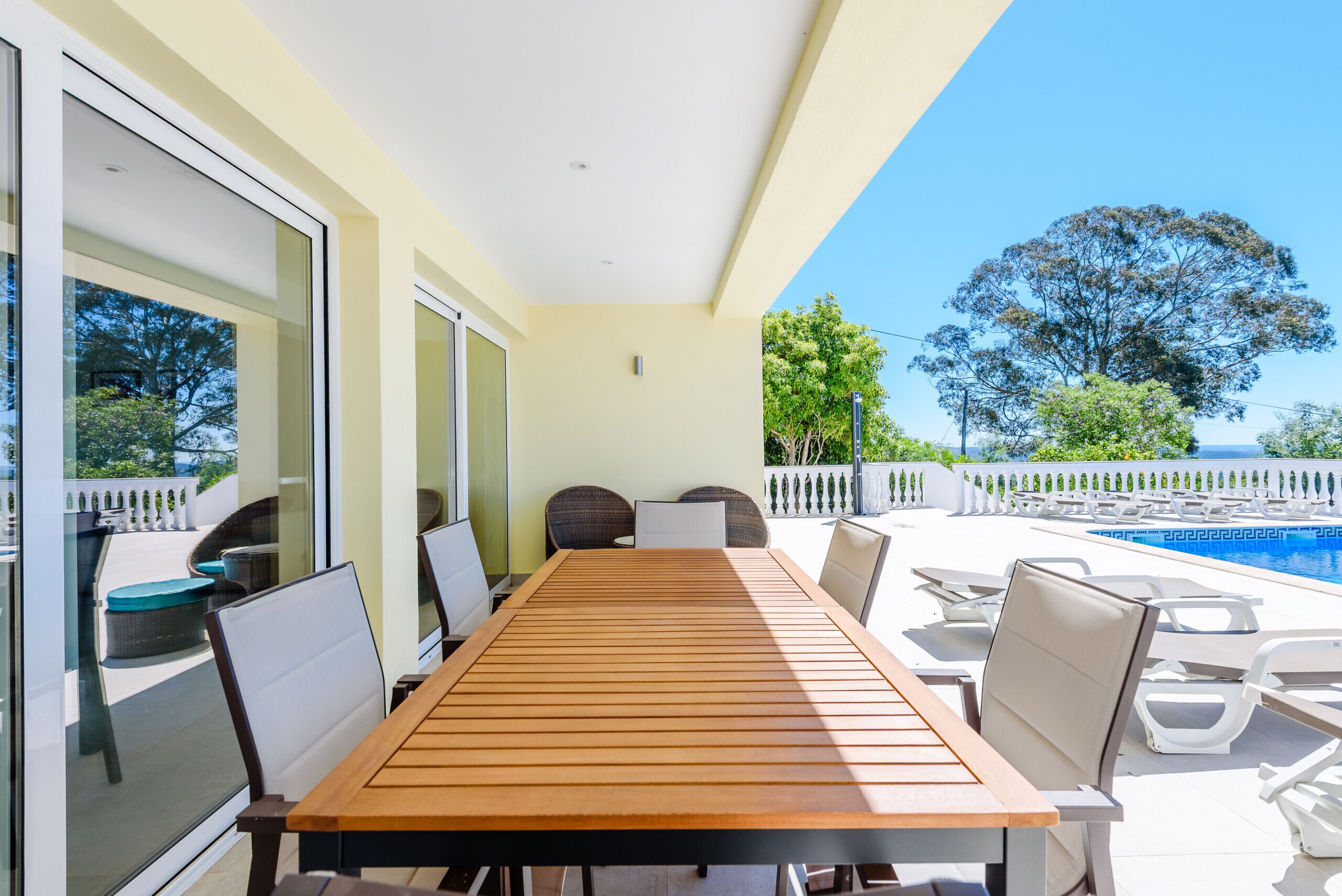 Un patio iluminado por el sol con una mesa de madera, sillas y vista a la piscina.