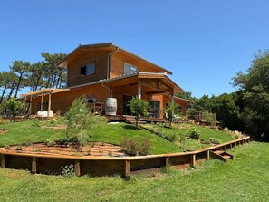 Ein zweistöckiges Holzhaus mit überdachter Veranda, einer kleinen Terrasse und einem angelegten Garten mit kreisförmigem Blumenbeet.