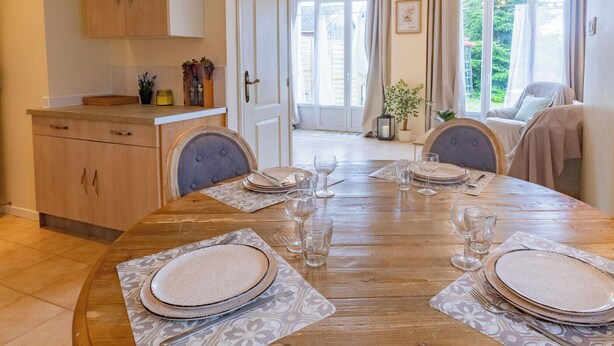 A dining area with a wooden table set for four, a kitchen with wooden cabinets, and a view of the outdoors through large windows.