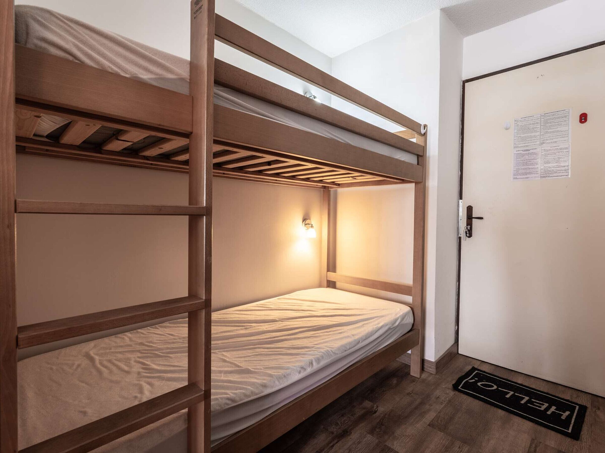 A bunk bed room with a wooden frame, a single bed, a nightstand with a lamp, a door with a doormat, and a wall with a notice.