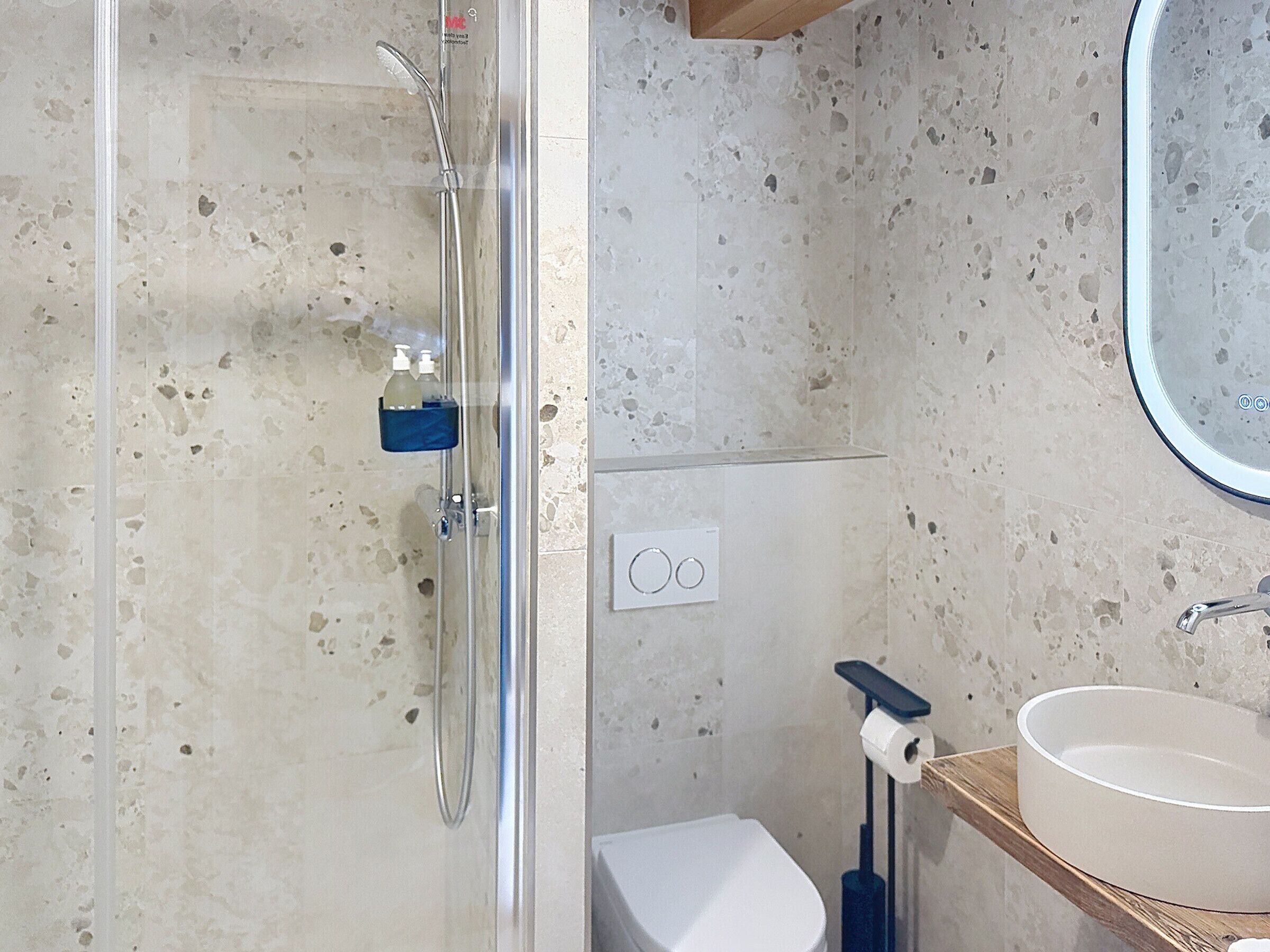 A bathroom with a glass-enclosed shower, a toilet, a sink with a mirror, and a soap dispenser on the wall.