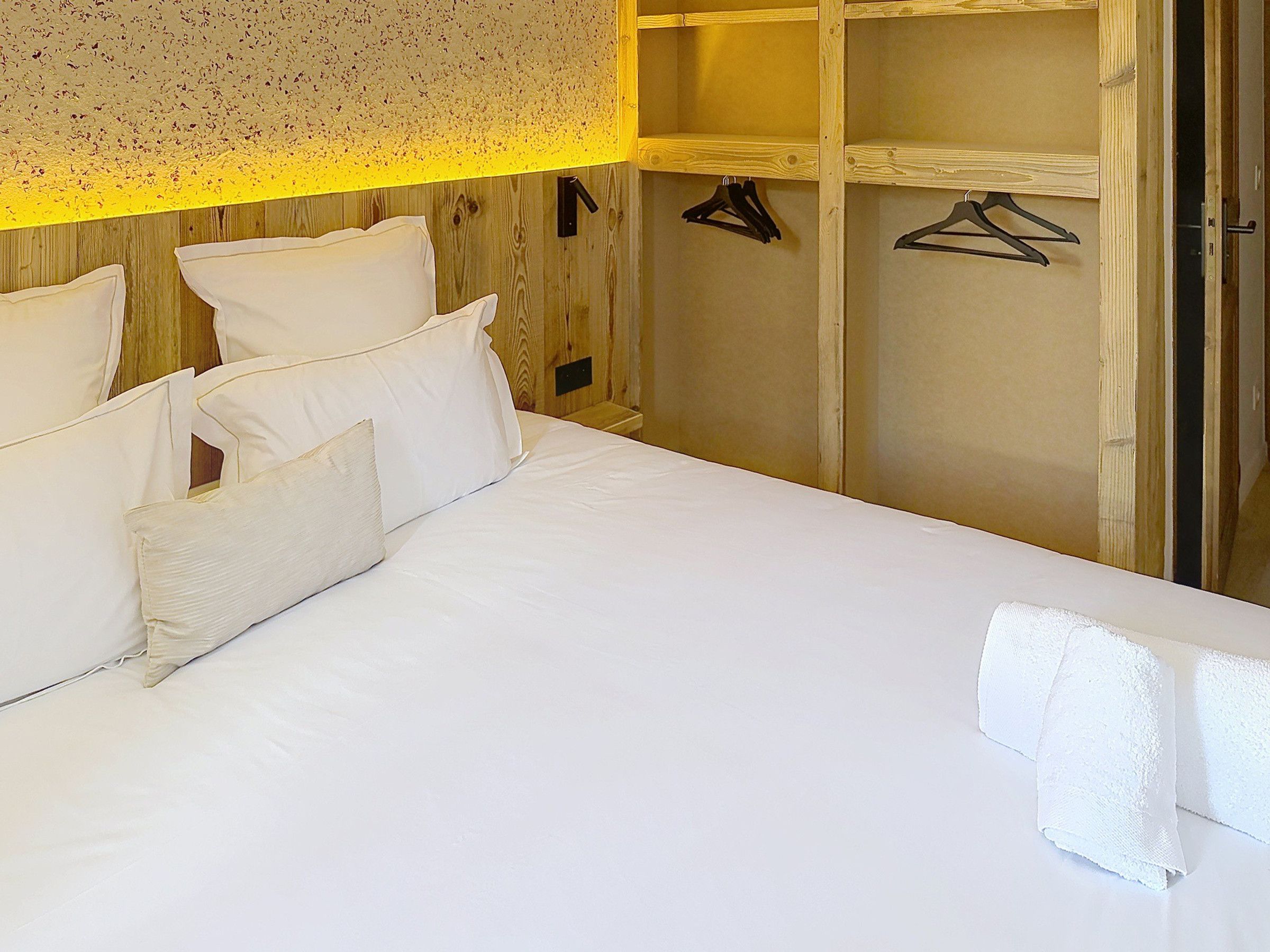Een keurig opgemaakt bed met witte lakens, een houten hoofdbord en een houten garderobe met kledinghangers.