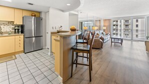 Una cocina moderna con una isla central, un comedor con una mesa redonda y sillas, y una sala de estar con sofá y balcón con vistas.