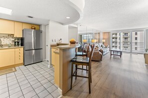 Eine moderne Küche mit Kochinsel, ein Essbereich mit rundem Tisch und Stühlen sowie ein Wohnzimmer mit Sofa und einem Balkon mit Aussicht.