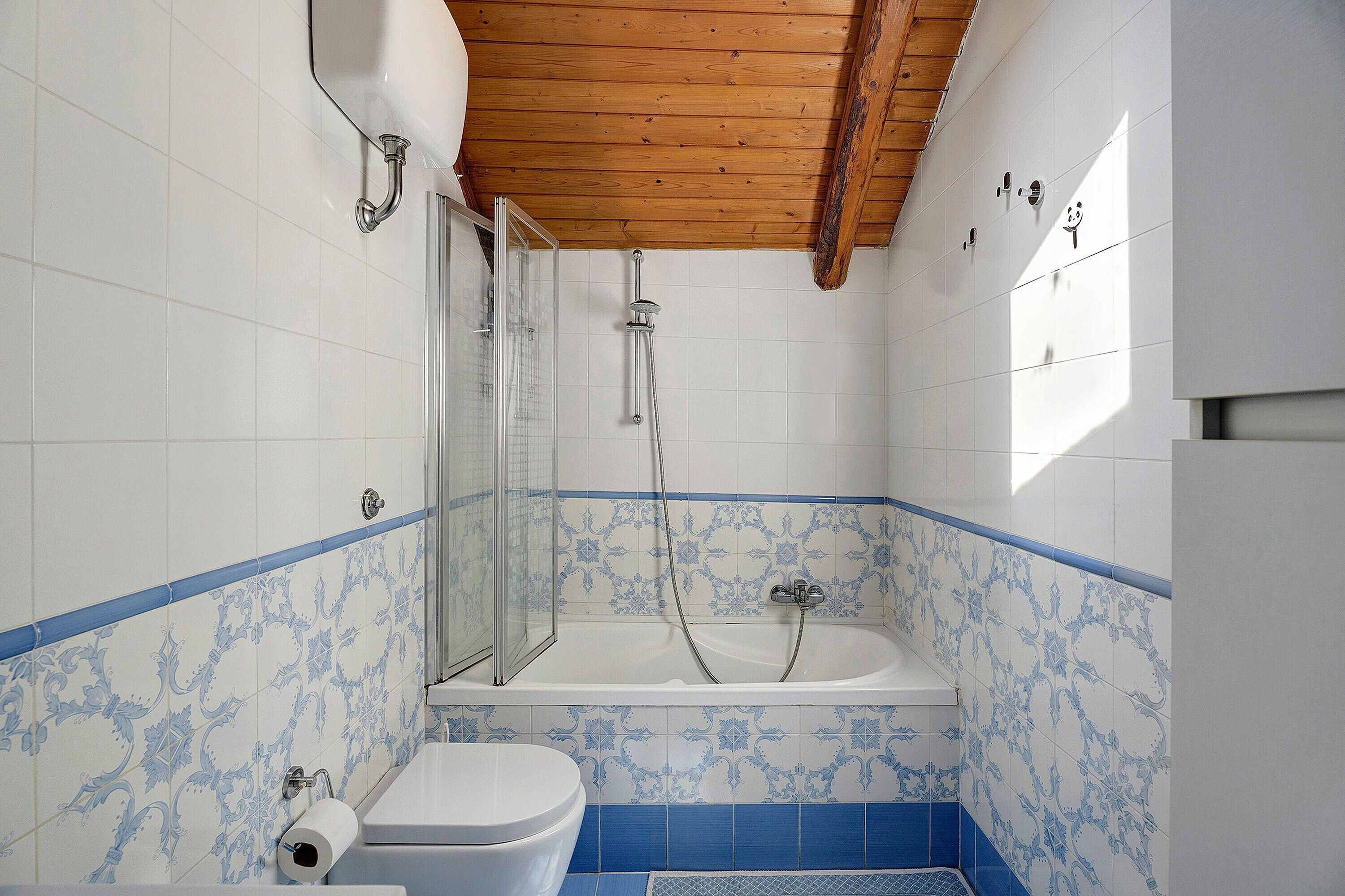 Een badkamer met douche, toilet en wastafel, voorzien van blauw-witte betegelde muren en houten balken in het plafond.