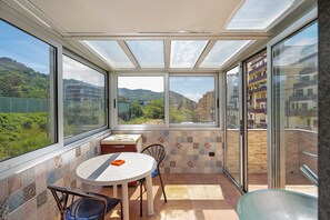 Een zonnig balkon met een tafel en stoelen, grote ramen en uitzicht op groen en gebouwen.