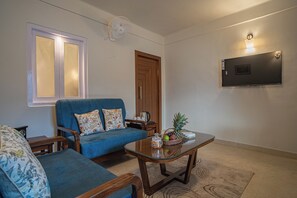 A living room with a blue sofa, a glass coffee table, a TV mounted on the wall, and a wooden door. - Alaya Stays Headingly Suite Lake Facing (Nainital)