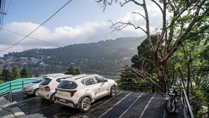 A parking area with two cars and a motorcycle, surrounded by trees and overlooking a hilly landscape. - Alaya Stays Headingly Suite Lake Facing (Nainital)
