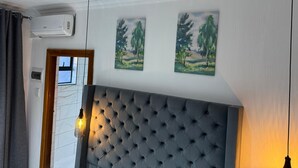 A room with a tufted headboard, two paintings of trees, an air conditioning unit, and a hanging light fixture. - Boutique Hotel-Style Room In Ferndale-A1 (Randburg)