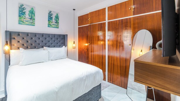 A modern bedroom with a tufted headboard, a wooden wardrobe, a desk with a mirror, and two wall-mounted paintings. - Boutique Hotel-Style Room In Ferndale-A1 (Randburg)