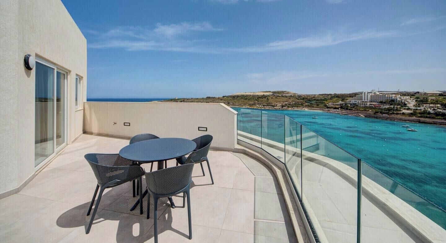 A balcony with a table and chairs, overlooking a clear blue sea and a distant landscape.