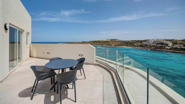 Un balcon avec une table et des chaises donnant sur une mer d’un bleu profond et sur une côte lointaine.