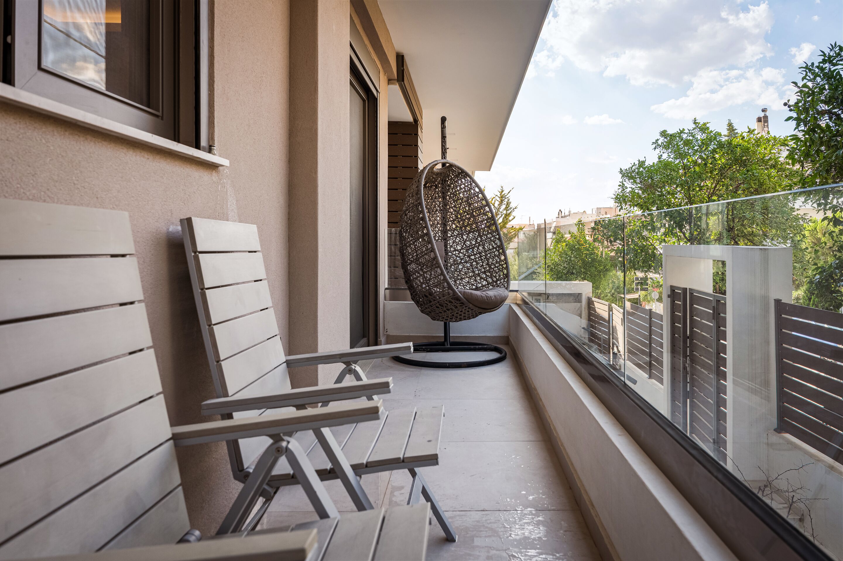 A balcony with a hanging chair, a bench, and a view of greenery and buildings.