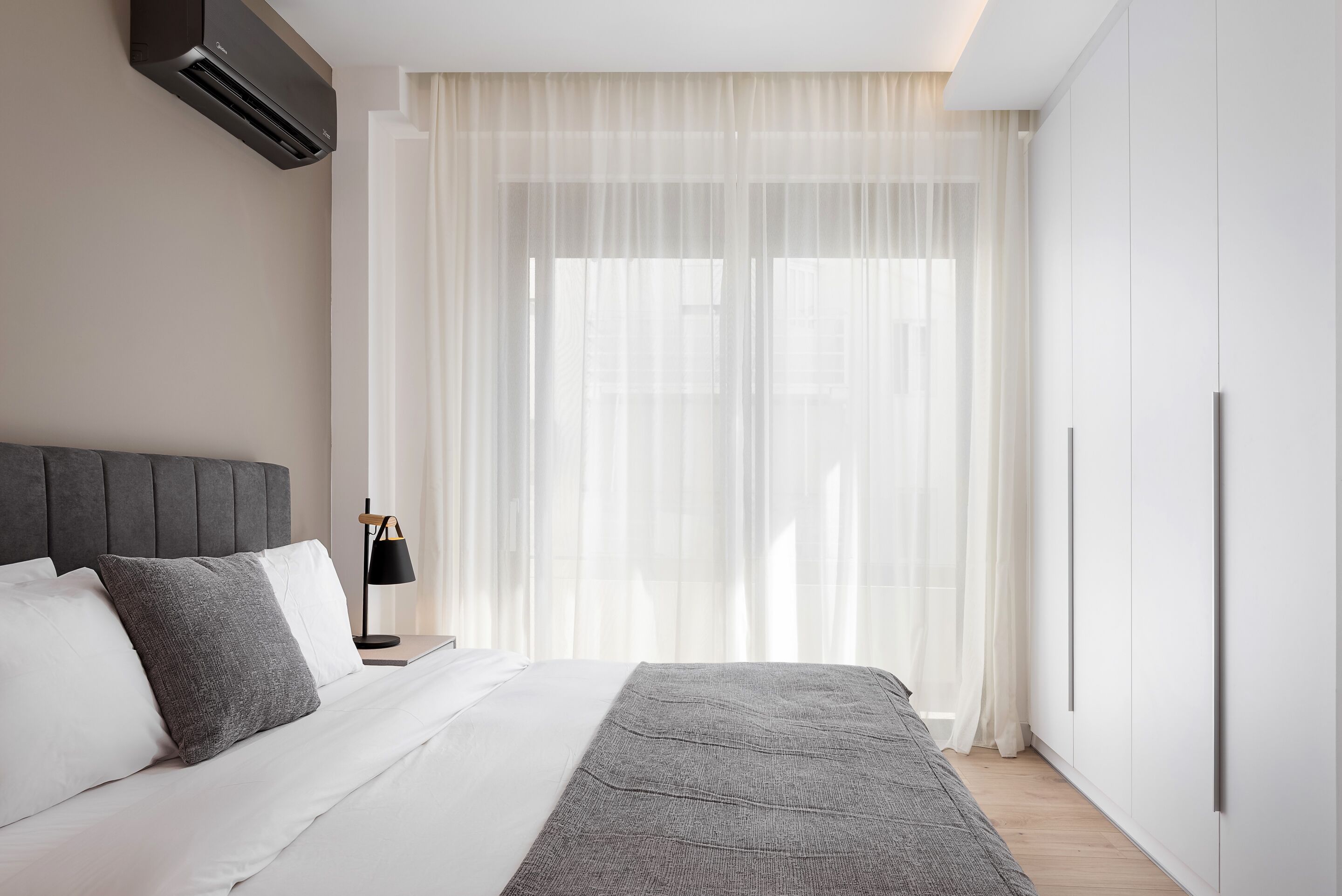 A modern bedroom with a bed, a bedside table, a lamp, and a large window with sheer curtains.