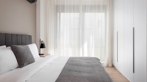 A modern bedroom with a bed, a bedside table, a lamp, and a large window with sheer curtains. - Alimos Escape 1 (Alimos)