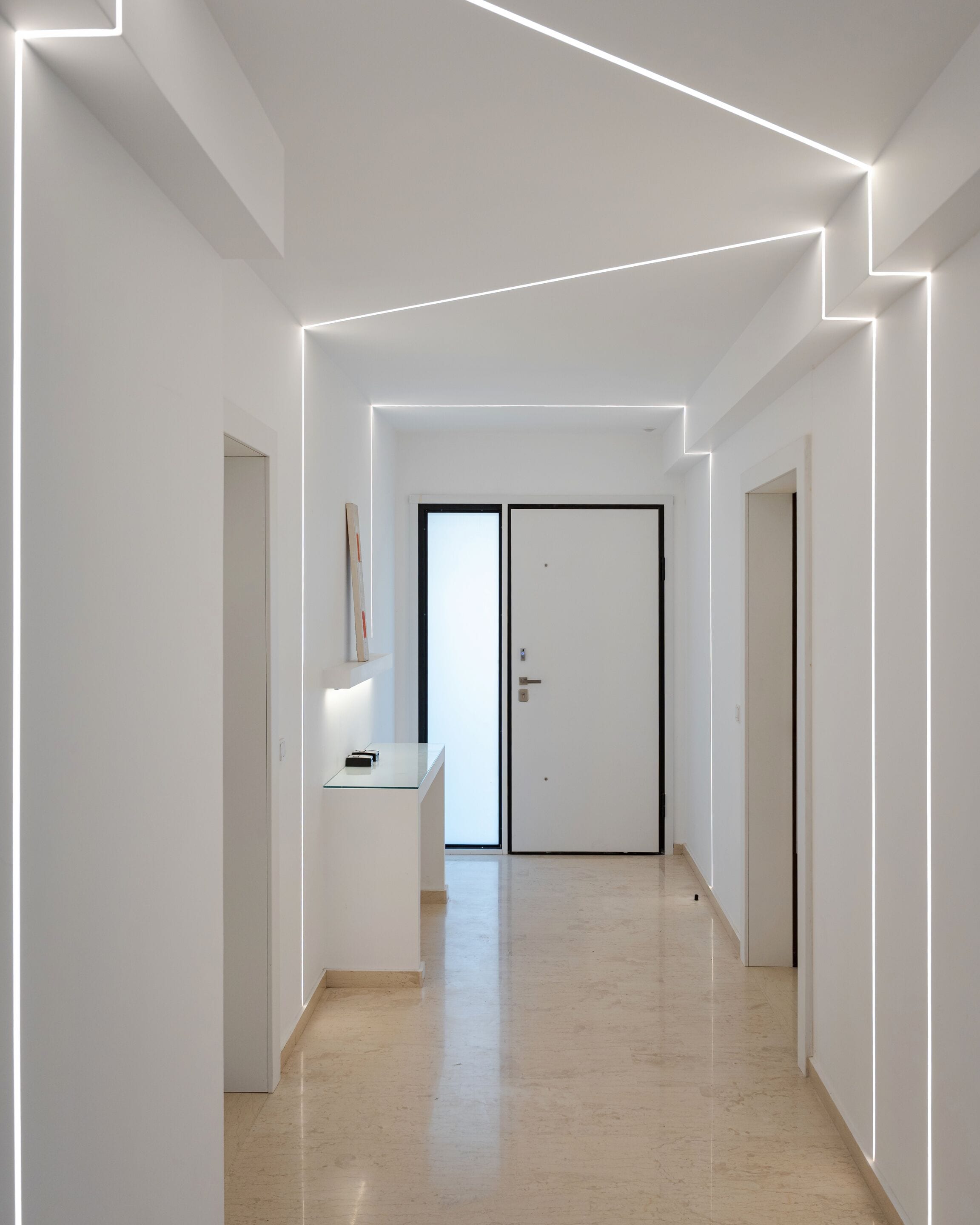 Un couloir moderne au design minimaliste, comprenant un bureau avec une lampe, une porte avec une poignée et une bande lumineuse au plafond.