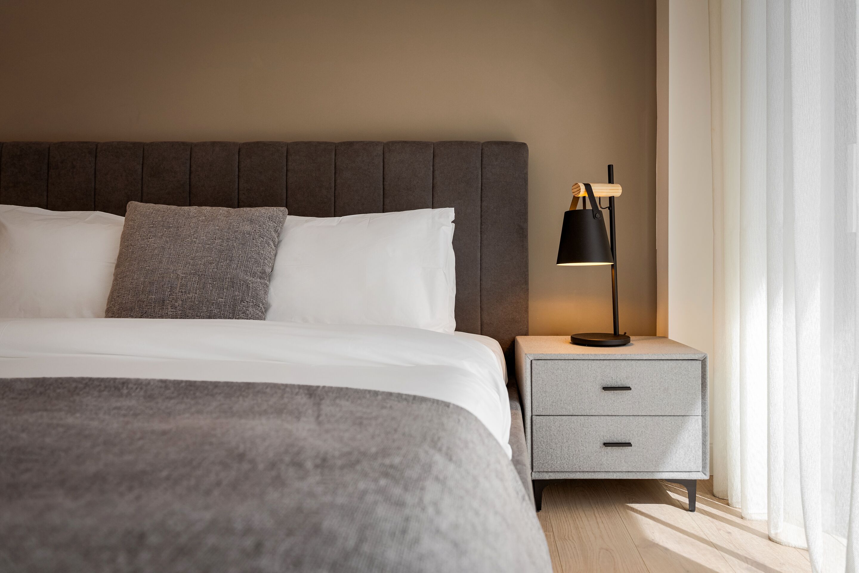 Una cama bien hecha con cabecera gris, una mesita de noche gris con una lámpara y una ventana con cortinas traslúcidas.