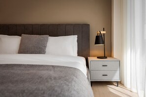 A neatly made bed with a grey headboard, a grey bedside table with a lamp, and a window with sheer curtains. - Alimos Escape 1 (Alimos)