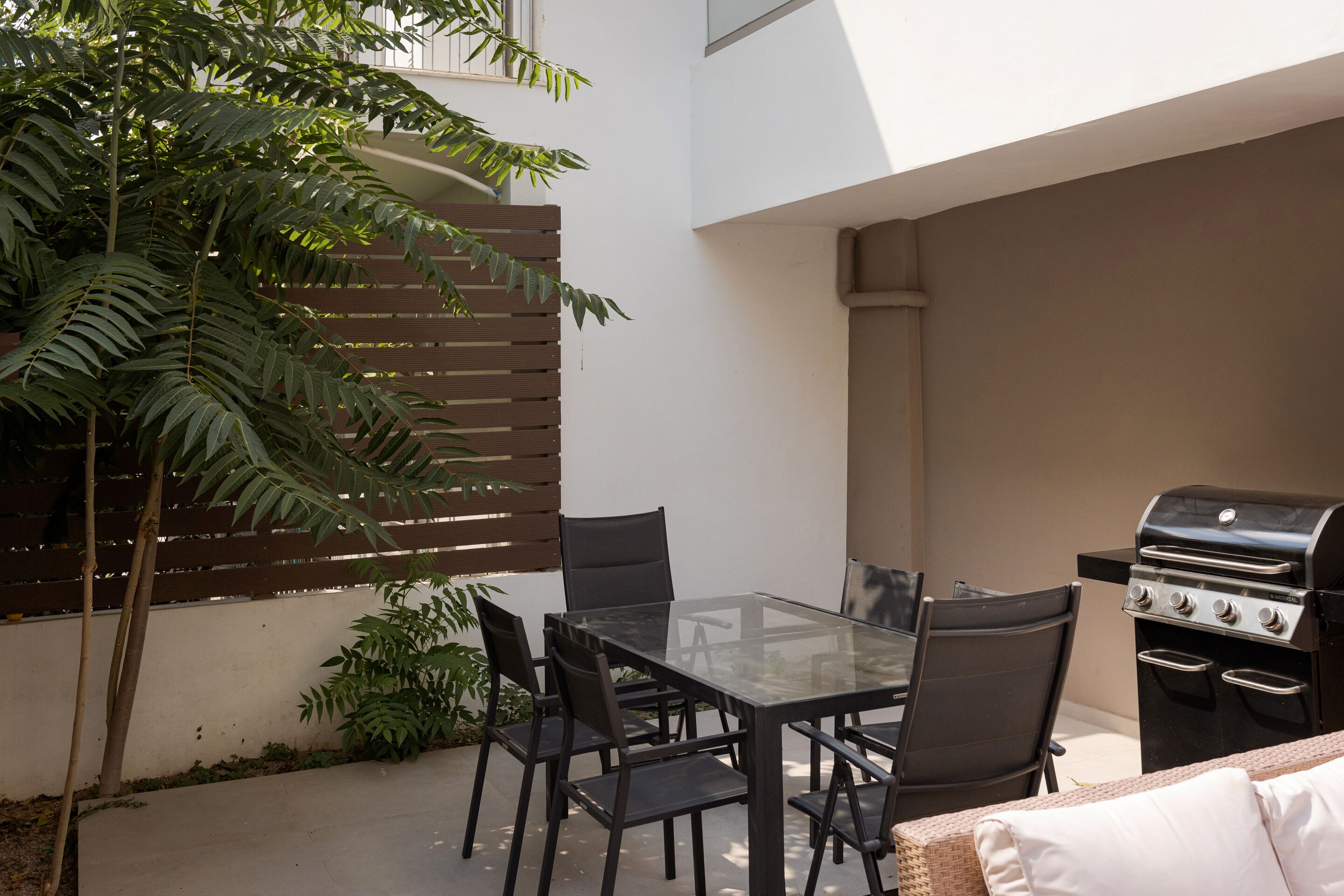 A patio with a glass table and chairs, a grill, and a potted plant.