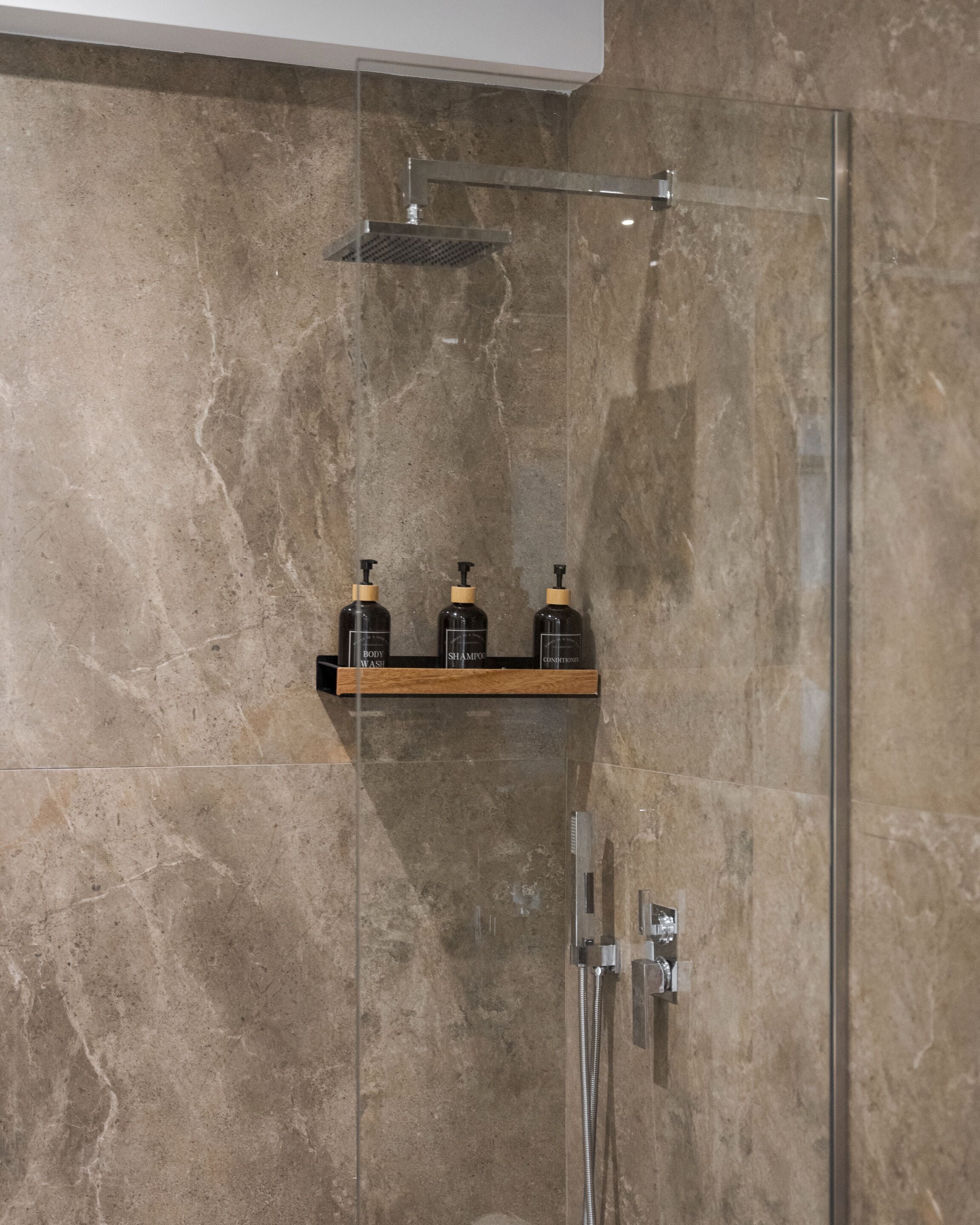 Un espace douche doté d’une porte en verre, d’un pommeau de douche à main et d’une étagère contenant trois bouteilles.