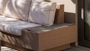 A wicker sofa with cushions against a wall, a glass door, and a concrete floor. - Alimos Escape 1 (Alimos)