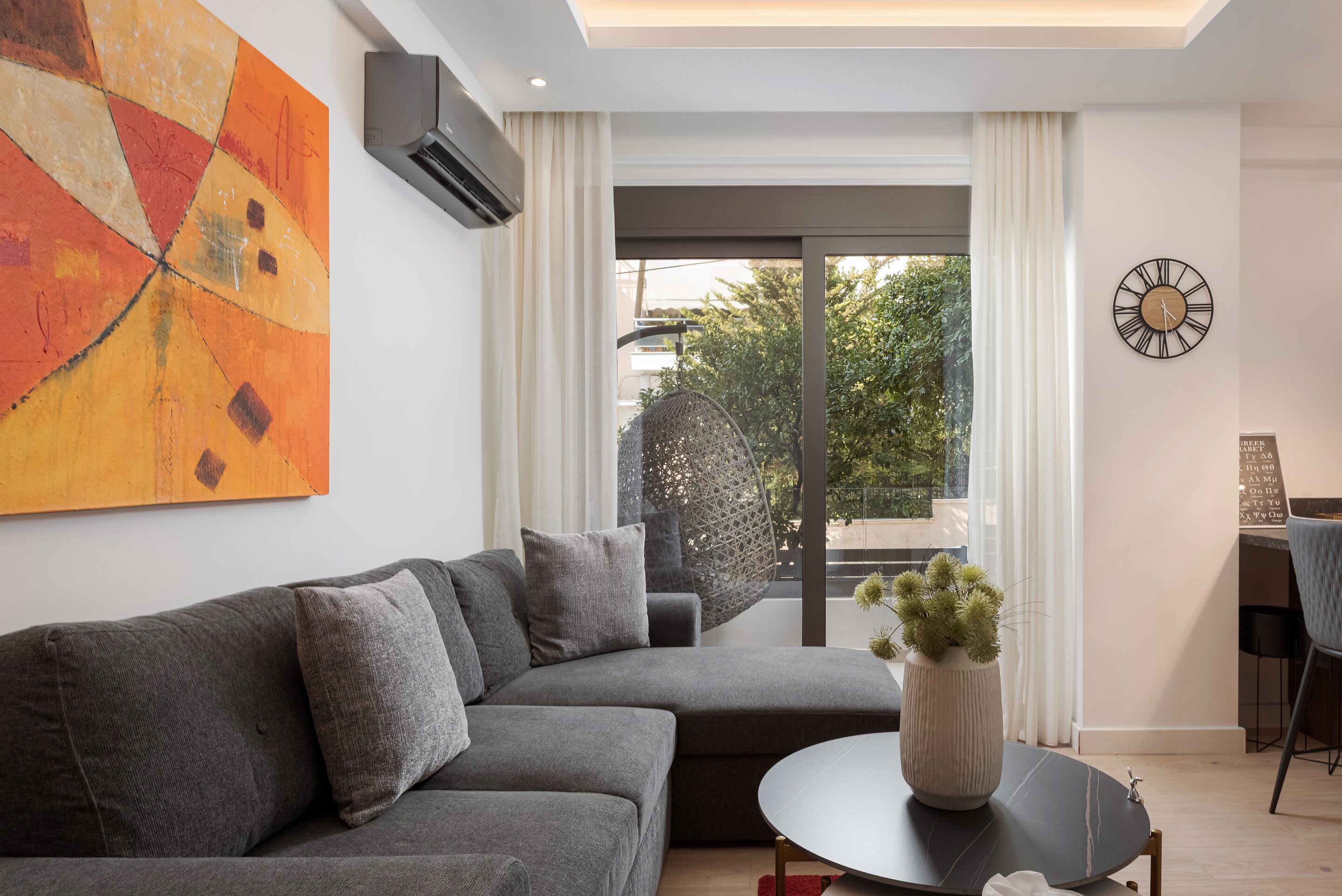 A modern living room with a sectional sofa, a round coffee table with a plant, a large abstract painting, and a view through a glass door.