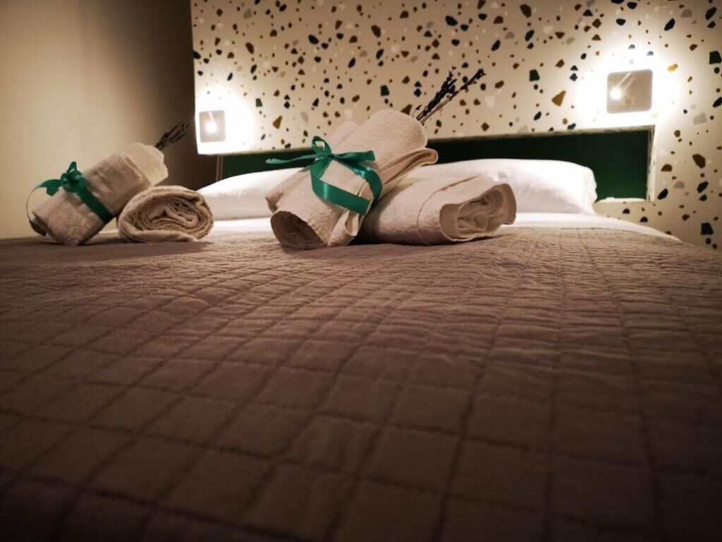 A neatly made bed with rolled towels and a green ribbon, a wall with a speckled pattern, and two wall-mounted lights.