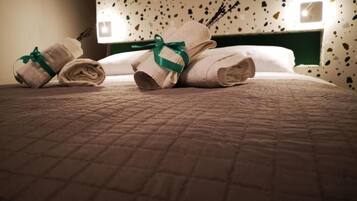Una cama bien tendida con toallas enrolladas y un lazo verde, una pared con un diseño moteado, y dos luces fijadas a la pared.