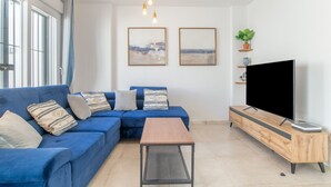 Een woonkamer met een blauwe bank, een houten salontafel, een televisie op een houten dressoir en twee ingelijste foto's aan de muur.