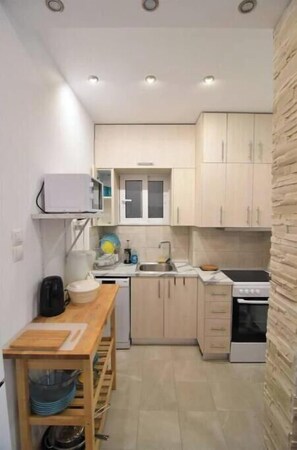 A compact kitchen with light-colored cabinets, a microwave, a sink, a stove, and a small dining table with a cutting board. - Modern 1-Bedroom Condo in Athens (Athina)