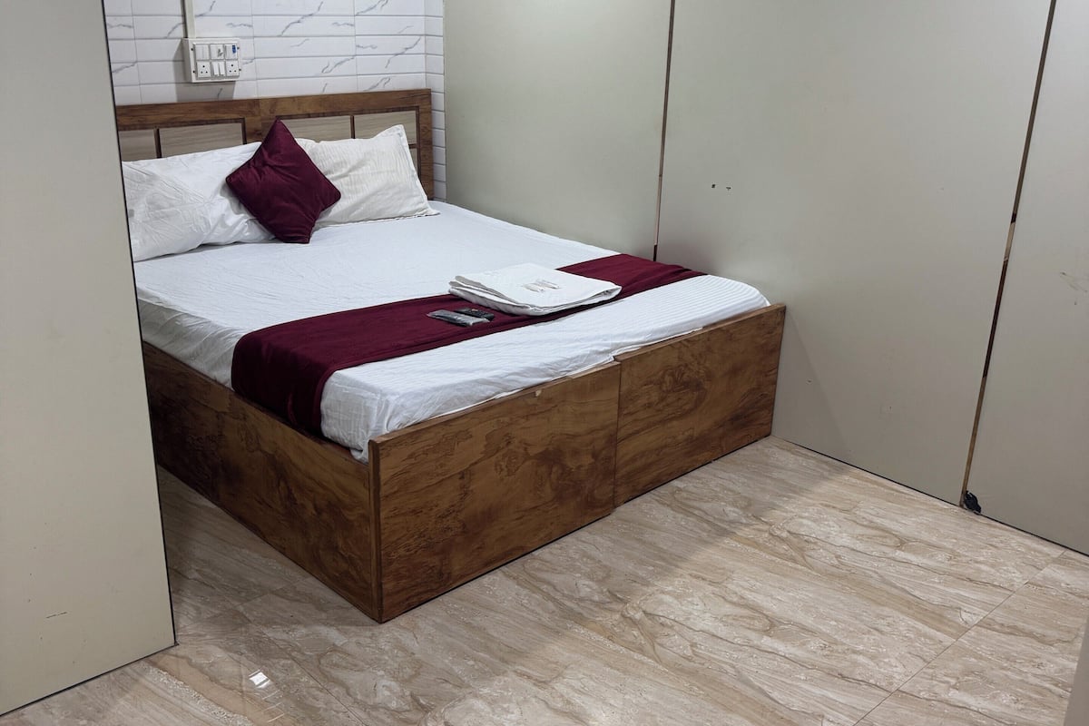 A bedroom with a wooden bed, white bedding, a marble-patterned floor, a ceiling fan, and a white air conditioning unit.