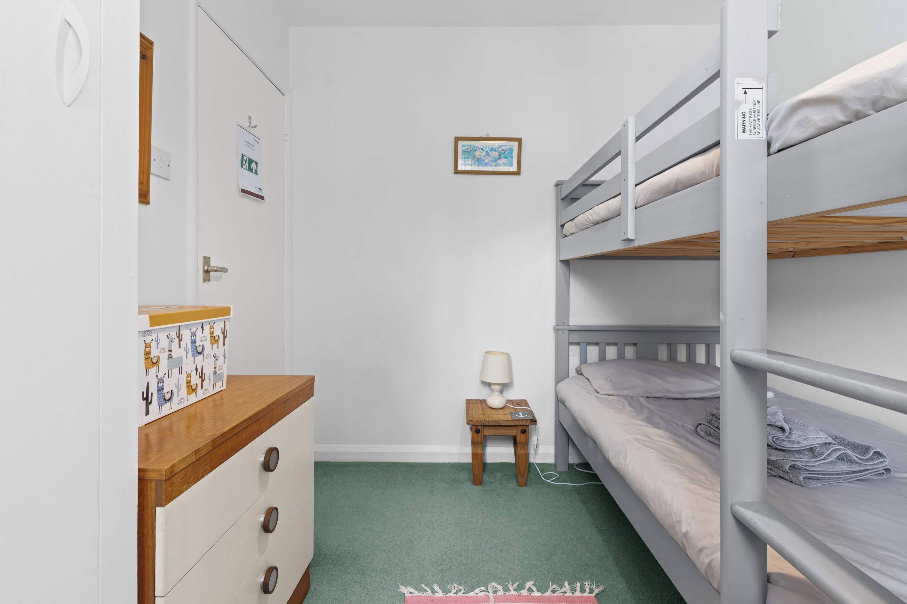 A bunk bed room with a wooden dresser, a small table with a lamp, and a framed picture on the wall.
