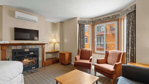 Ein Hotelzimmer mit Kamin, Fernseher, zwei Sesseln, einer Couch, einem kleinen Tisch und einem fenster mit Vorhängen.